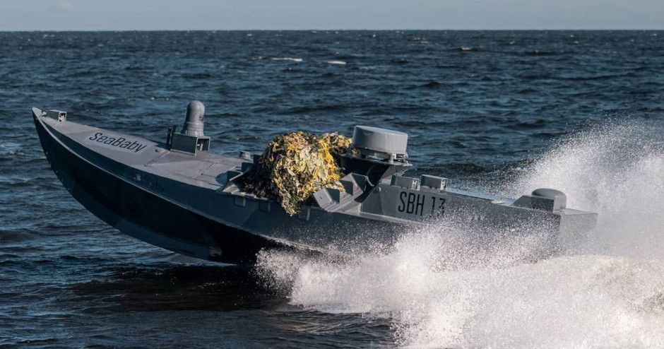 A military-style boat labeled SeaBaby SBH 13 speeds through choppy waters with a camouflage net on its deck. Waves splash up around the vessel against a backdrop of dark ocean water and a clear sky.
