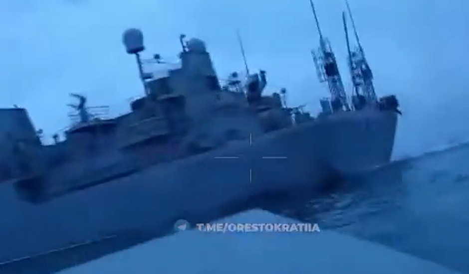 A large military ship with antennas and radar equipment navigates through the sea in overcast weather. The vessel appears tilted, and the photo has a blue tint.